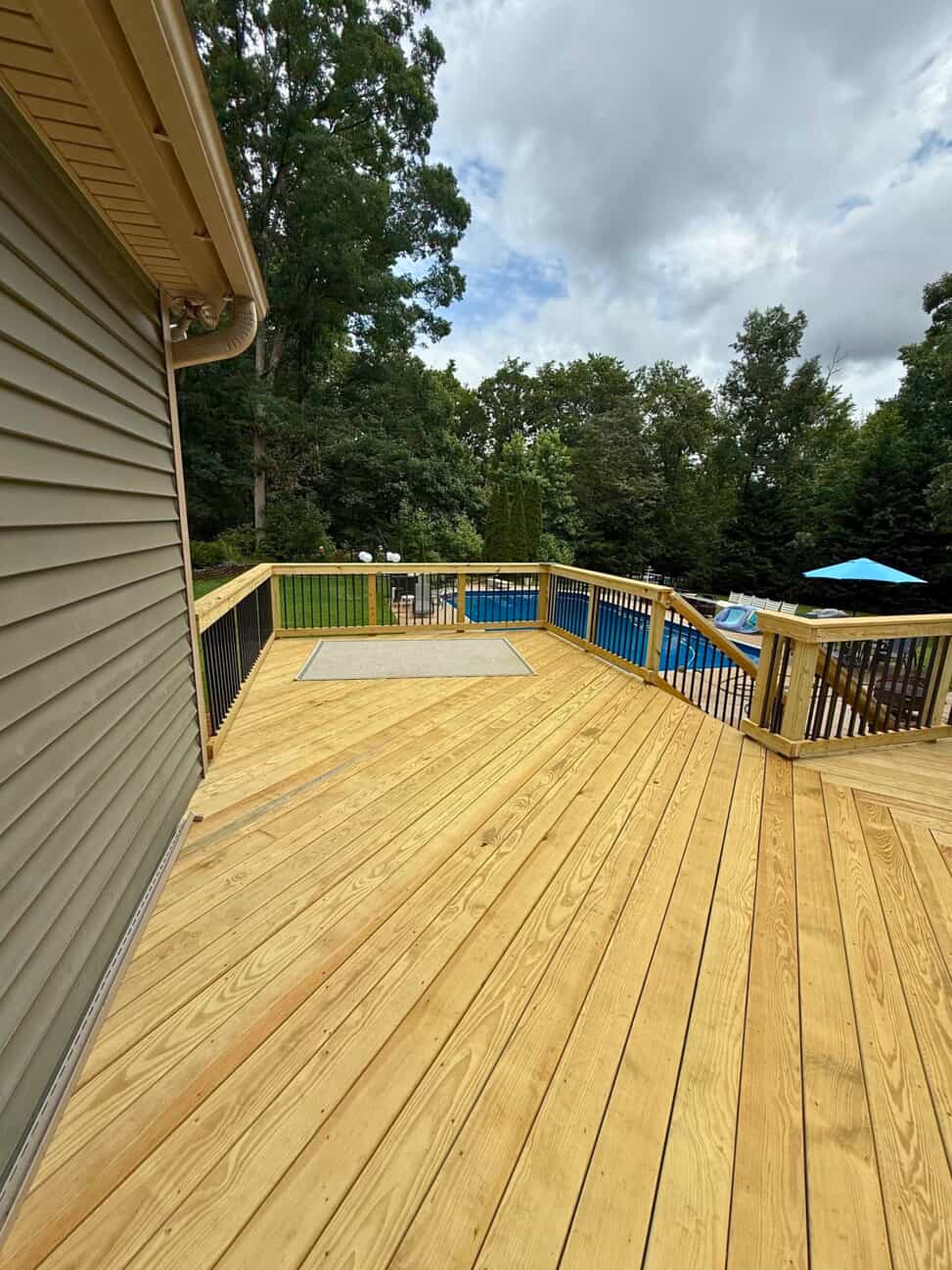 A wide, light-colored wood deck with black metal railings extending towards a backyard with an above-ground swimming pool. Lush green trees are visible in the background under a cloudy sky.